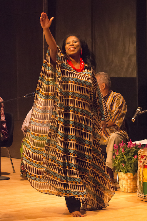 N'Deye Gueye wears a floor skimming brightly patterned dress and lifts one arm out above her